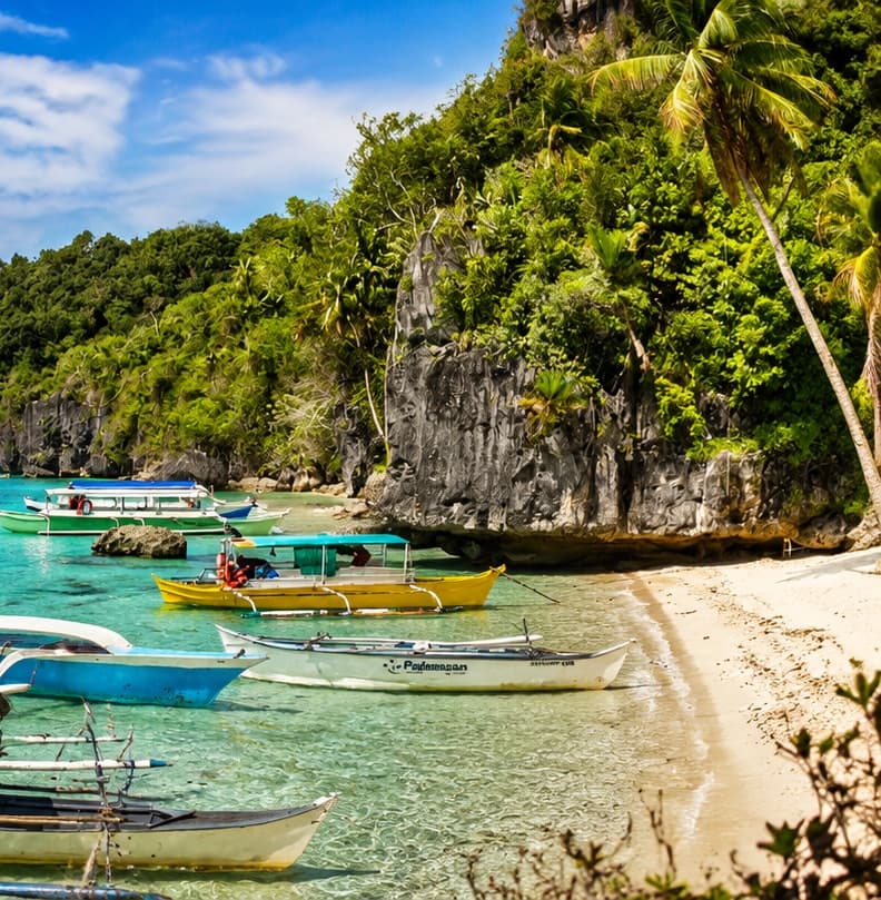 Outrigger bangka boats moored on clear shallow water beside a limestone cliff and white sand
