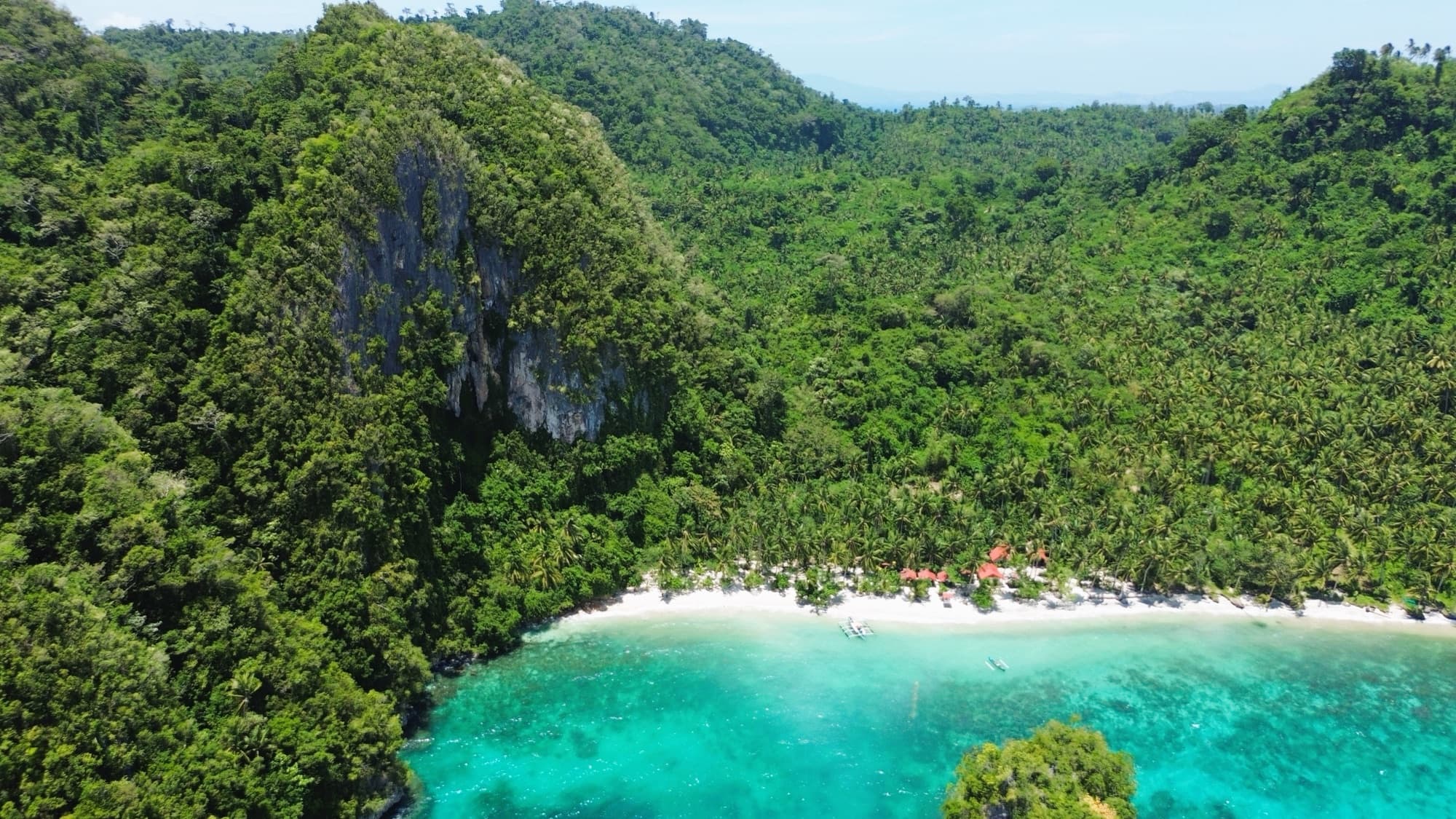 Aerial view of a forested limestone cove with a white sand beach and turquoise reef flats at Cabacungan Cove, Basilisa