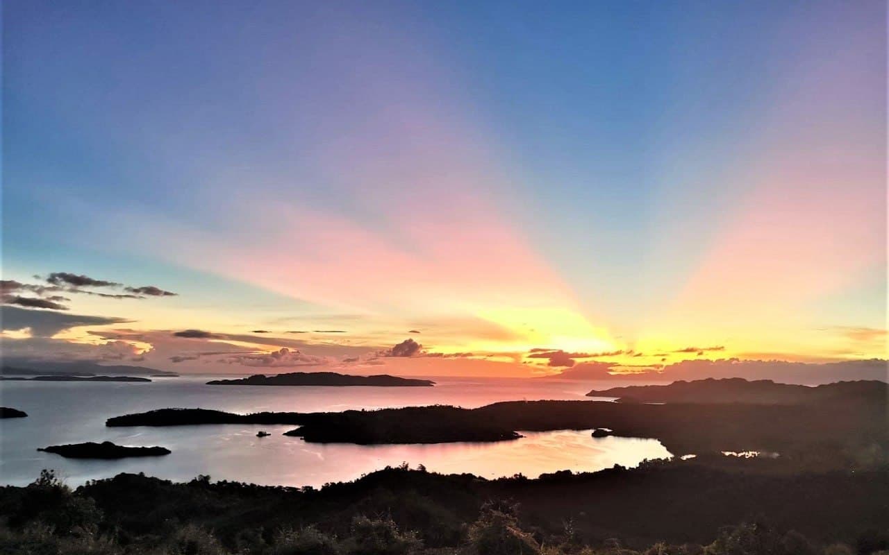 Sun rays fanning across a pink and blue sky over the islands, seen from Laaganan Skybar